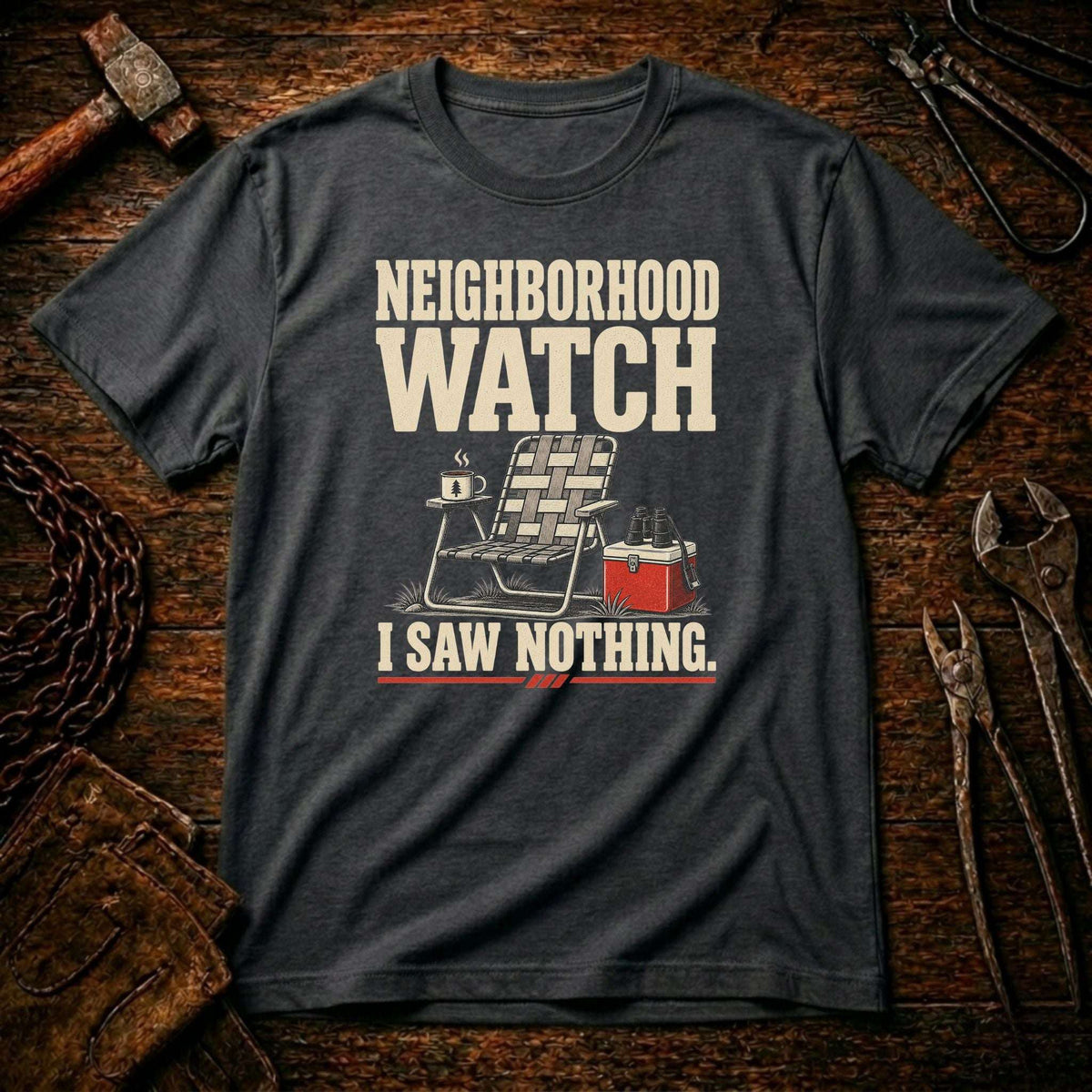 A dark charcoal gray graphic t-shirt laid flat on a rustic wooden background with tools like a hammer, chain, and wrenches. The shirt features a cream-colored graphic that reads 'NEIGHBORHOOD WATCH' above a folding chair with a mug and a red cooler, followed by 'I SAW NOTHING.