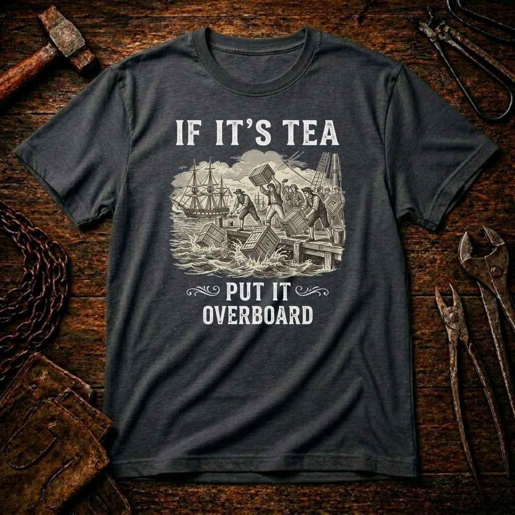 A dark charcoal gray graphic t-shirt laid flat on a rustic wooden background with tools like a hammer, chain, and wrenches. The shirt features a white vintage-style illustration of a shipwreck with the text 'IF IT'S TEA PUT IT OVERBOARD'.