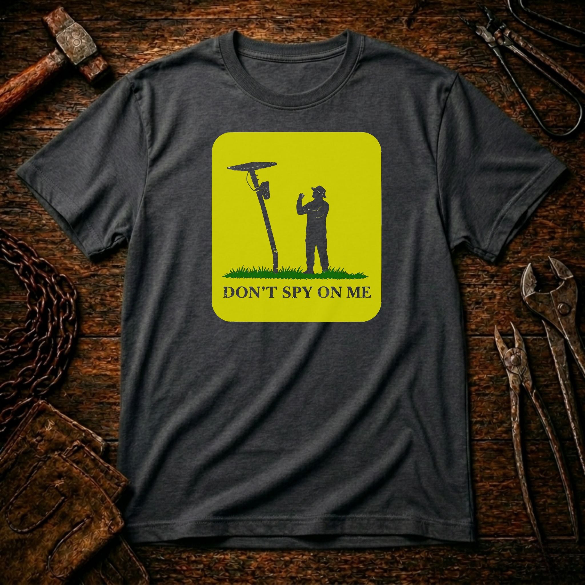 A dark charcoal gray t-shirt laid flat on a rustic wooden background with tools like a hammer, wrenches, and chain visible around it. The shirt features a bright yellow-green square graphic with a black silhouette of a person next to a spy radio antenna and the text 'DON'T SPY ON ME'.