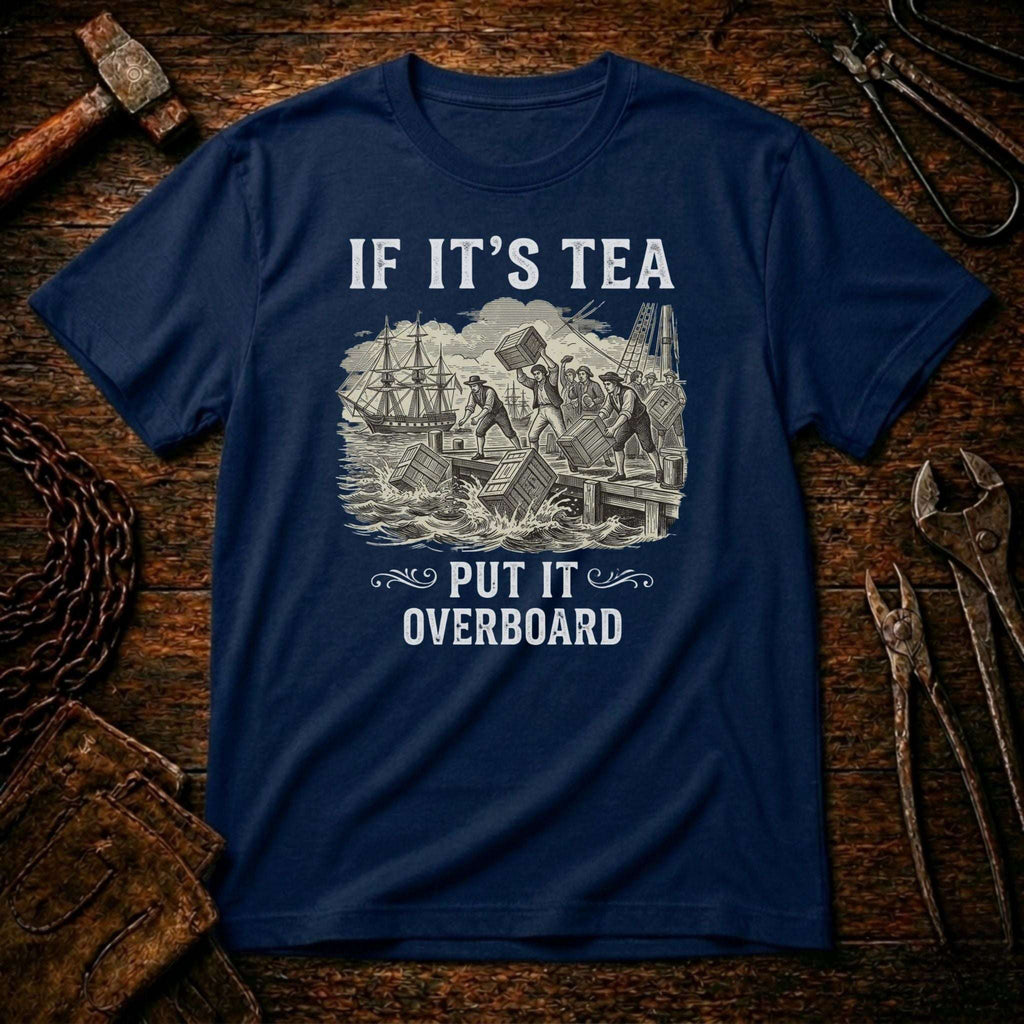 A navy blue crewneck t-shirt with a white graphic print reading 'IF IT'S TEA PUT IT OVERBOARD' and showing a vintage illustration of a shipwreck, displayed flat on a rustic wooden background with tools.
