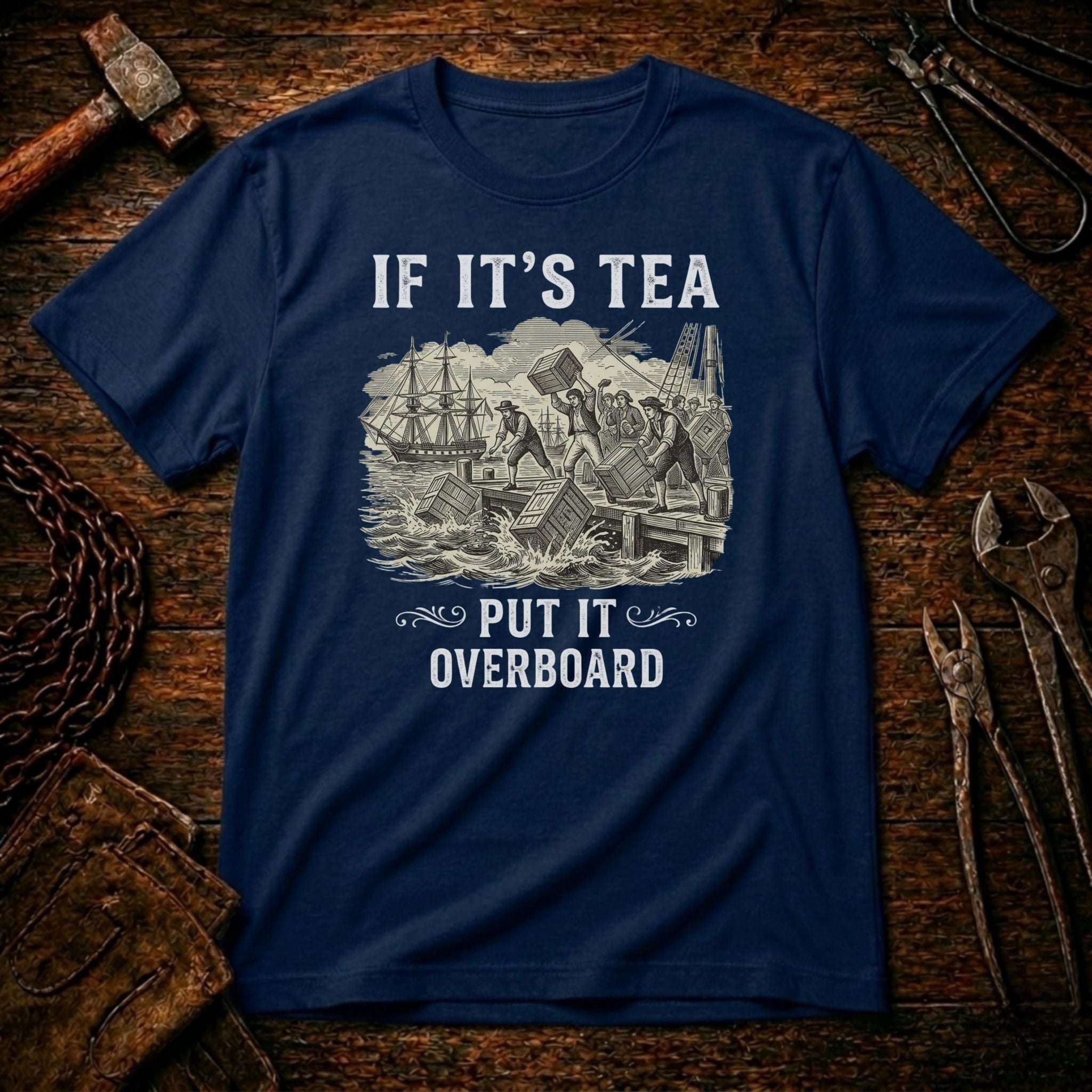 A navy blue crewneck t-shirt with a white graphic print reading 'IF IT'S TEA PUT IT OVERBOARD' and showing a vintage illustration of a shipwreck, displayed flat on a rustic wooden background with tools.