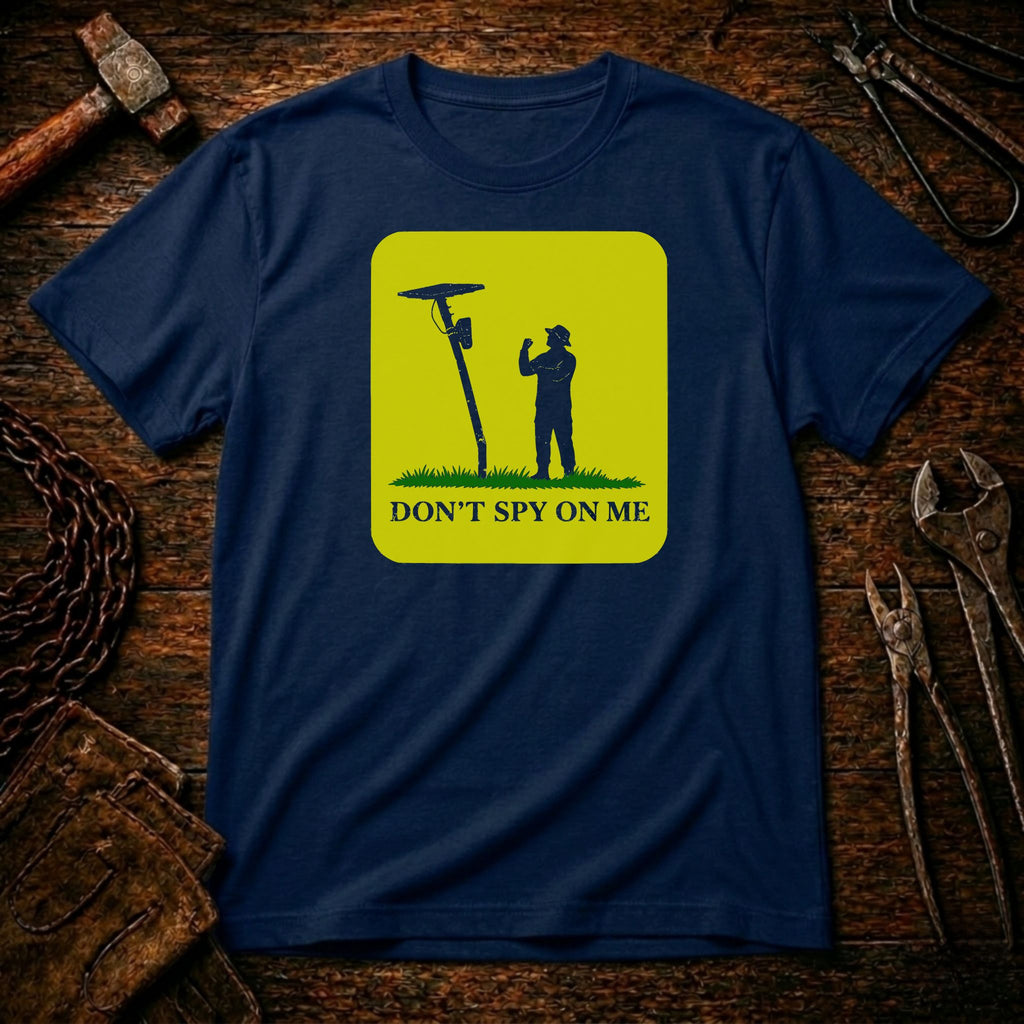 A navy blue crewneck t-shirt with a yellow-green graphic print showing a silhouette of a person next to a spy device and the text 'DON'T SPY ON ME'. The shirt is displayed flat on a rustic wooden background surrounded by tools like a hammer, wrenches, and a chain.