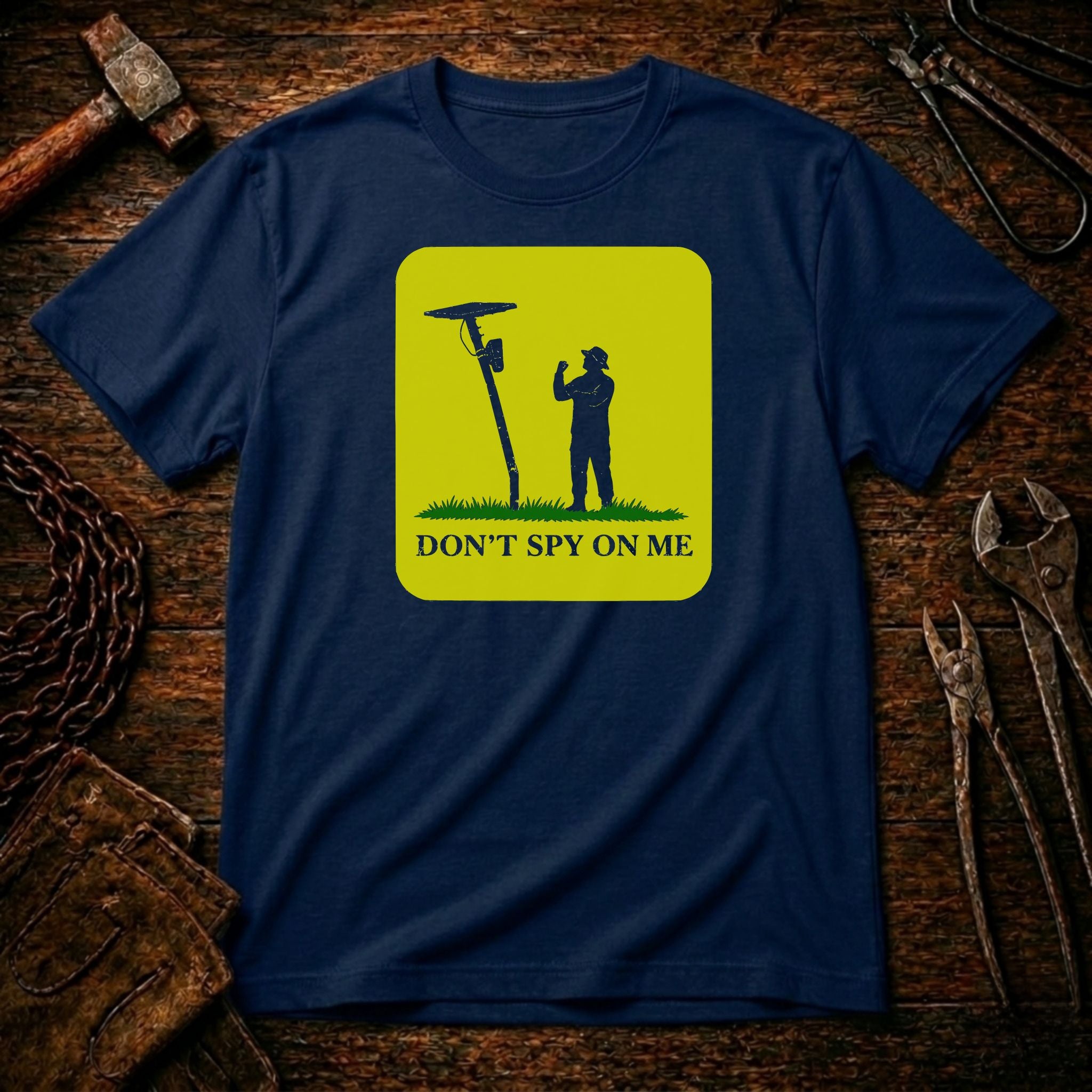 A navy blue crewneck t-shirt with a yellow-green graphic print showing a silhouette of a person next to a spy device and the text 'DON'T SPY ON ME'. The shirt is displayed flat on a rustic wooden background surrounded by tools like a hammer, wrenches, and a chain.
