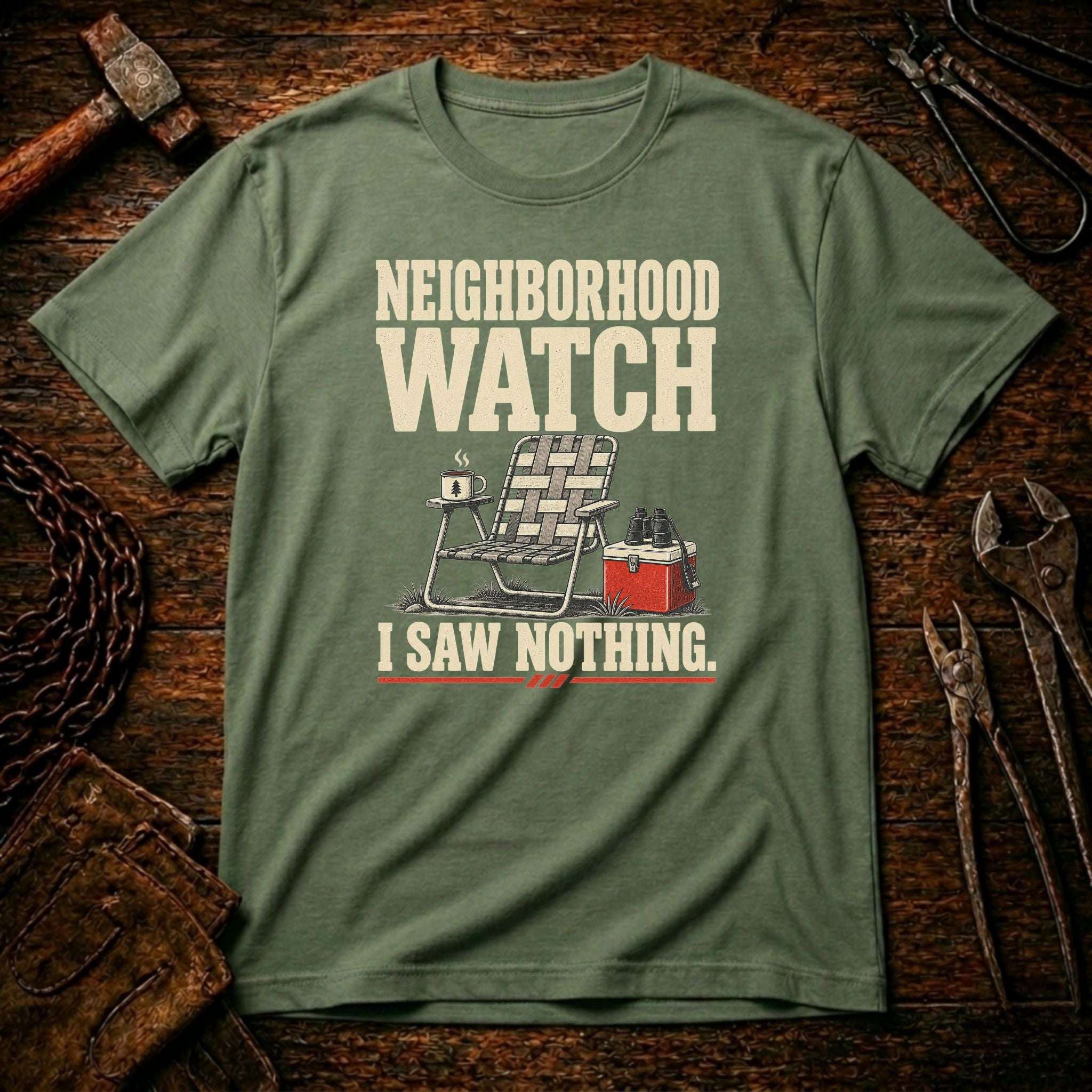 A military green graphic t-shirt laid flat on a rustic wooden background with tools like a hammer, chain, and wrenches. The shirt features a cream-colored graphic that reads 'NEIGHBORHOOD WATCH I SAW NOTHING.