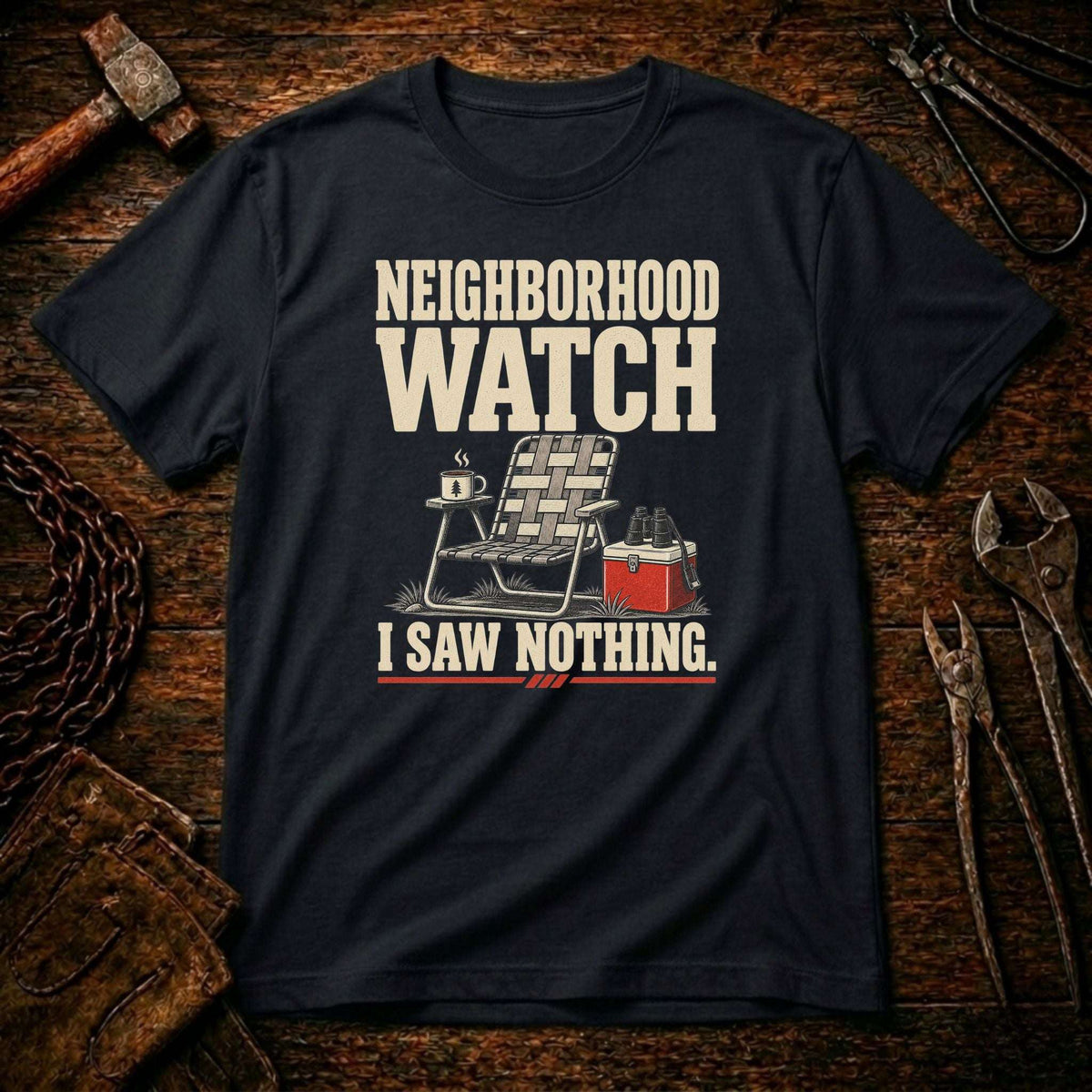 A black graphic t-shirt laid flat on a rustic wooden background with tools like a hammer, chain, and wrenches. The shirt features a graphic print of a folding chair, a mug, and a red cooler with the text 'NEIGHBORHOOD WATCH I SAW NOTHING.