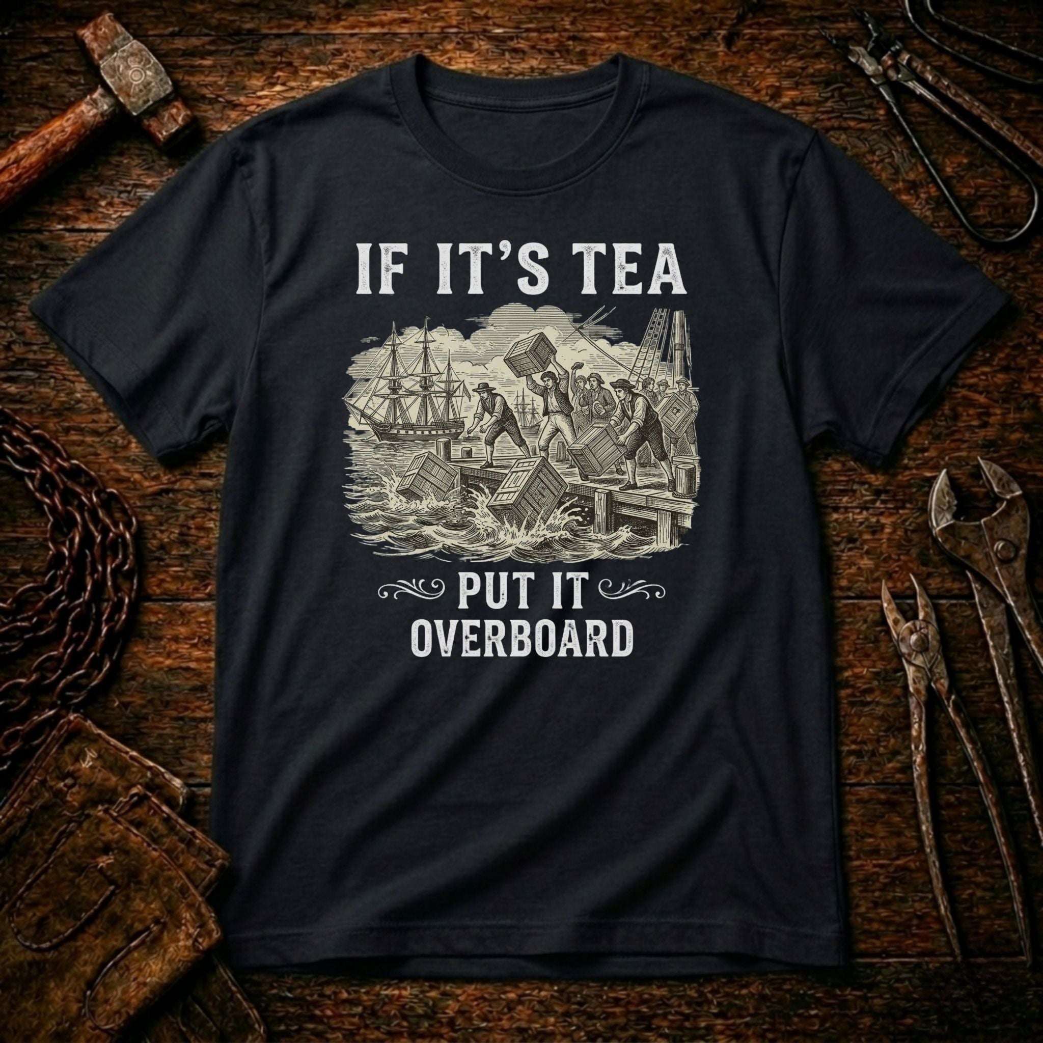 A black crewneck t-shirt with a vintage-style nautical graphic and the text 'IF IT'S TEA PUT IT OVERBOARD', displayed flat on a rustic wooden background with tools.