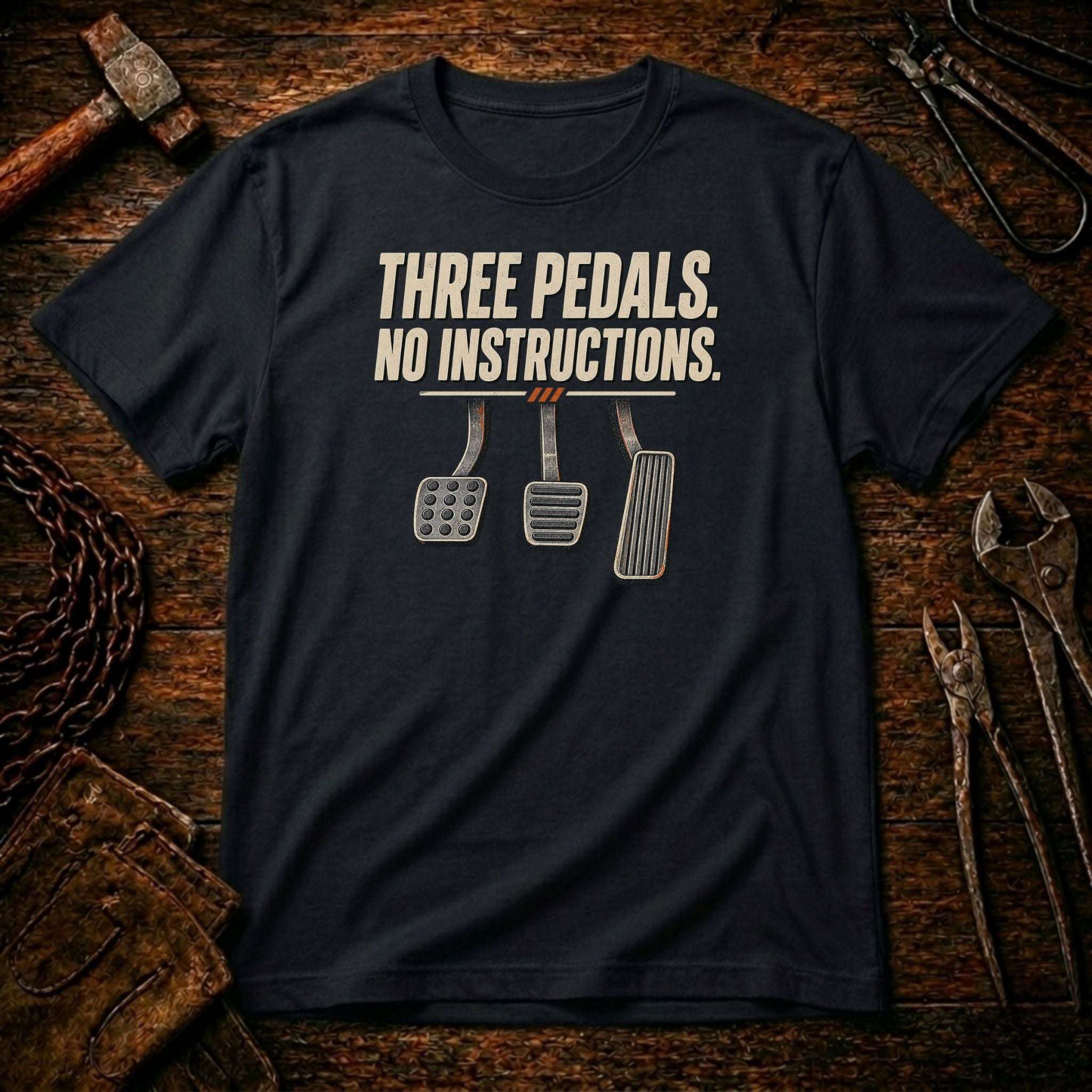 A black graphic t-shirt laid flat on a rustic wooden workbench surrounded by tools. The shirt features a graphic of three car pedals (clutch, brake, accelerator) with the text 'THREE PEDALS.