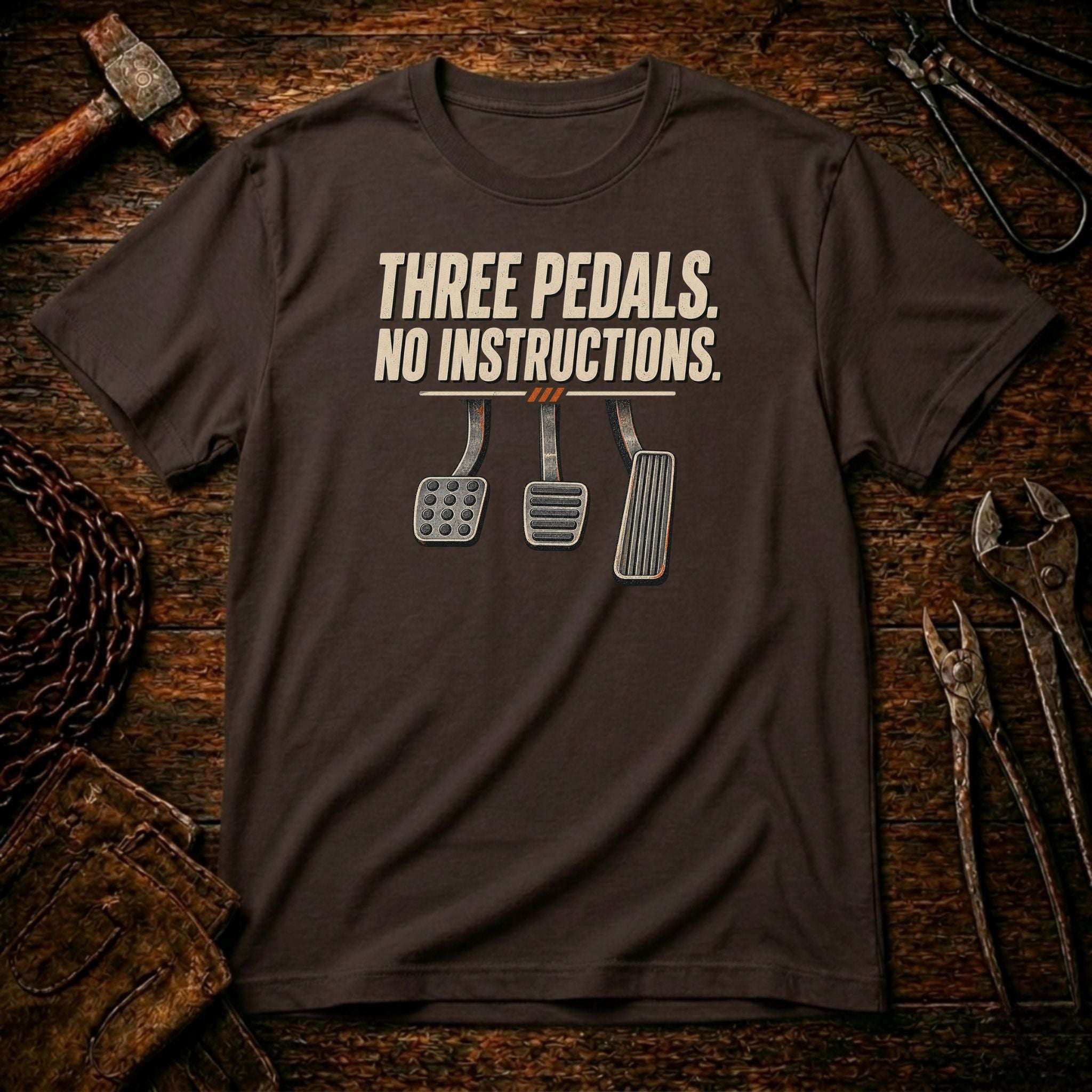 A dark brown t-shirt with a graphic of three car pedals and the text 'THREE PEDALS. NO INSTRUCTIONS.