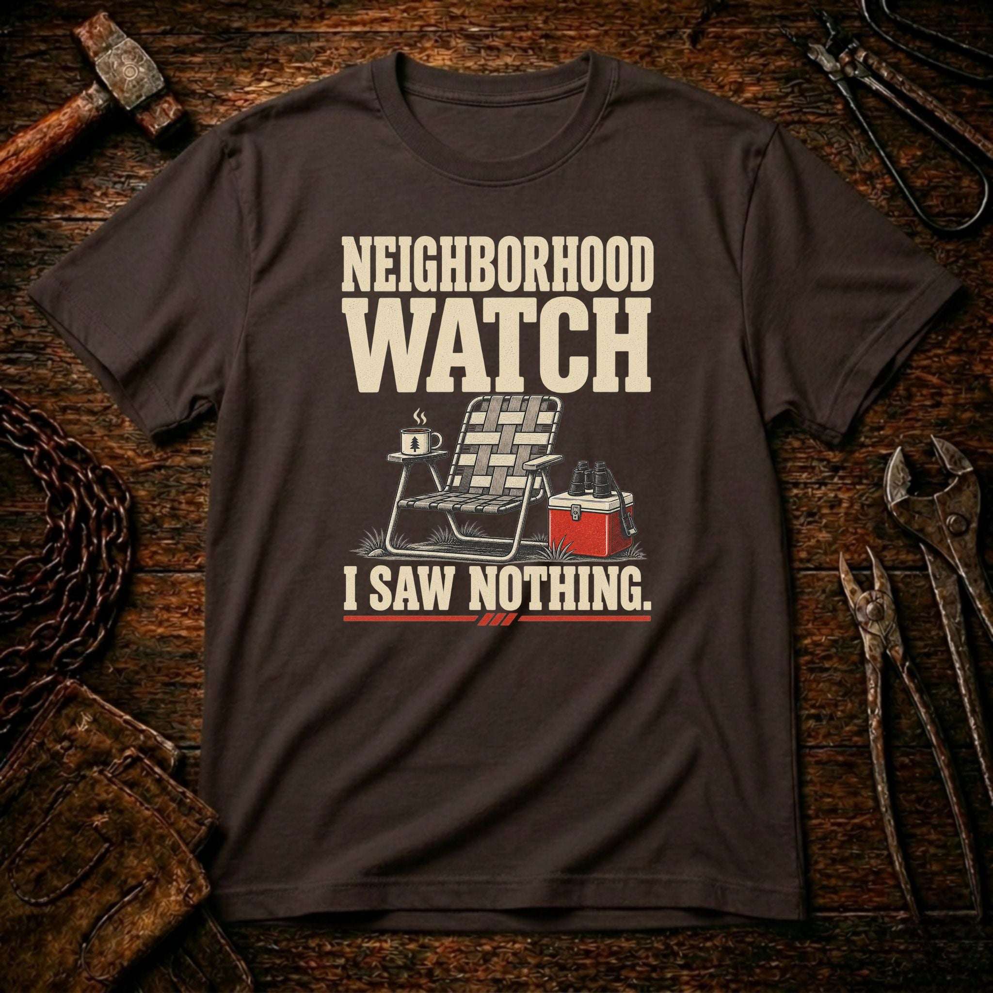 A dark brown graphic t-shirt laid flat on a rustic wooden surface surrounded by tools like a hammer, chain, and wrenches. The shirt features a graphic of a folding chair and a red cooler with the text 'NEIGHBORHOOD WATCH I SAW NOTHING.