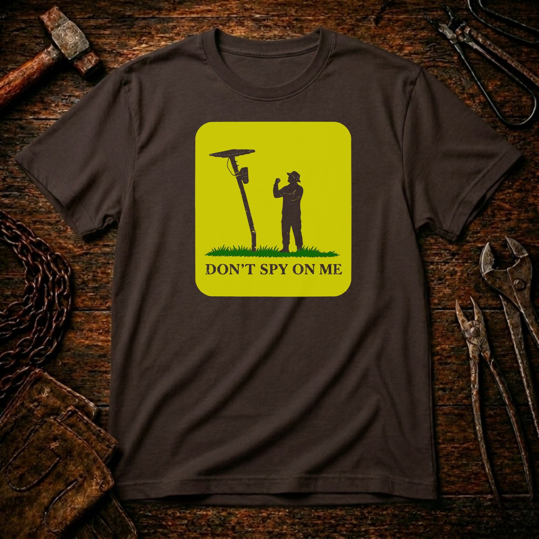 A dark brown t-shirt laid flat on a rustic wooden background with tools like a hammer, wrenches, and pliers. The shirt features a bright yellow-green square graphic with a black silhouette of a person holding a spy camera next to a satellite dish, with the text 'DON'T SPY ON ME' below.