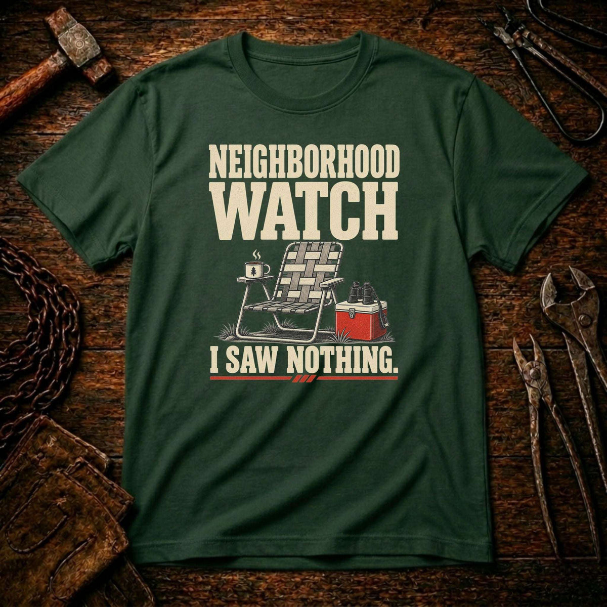 A forest green graphic t-shirt laid flat on a rustic wooden background with tools like a hammer, chain, and wrenches. The shirt features a cream-colored graphic that reads 'NEIGHBORHOOD WATCH' above a folding chair, a mug, and a red cooler, with 'I SAW NOTHING.
