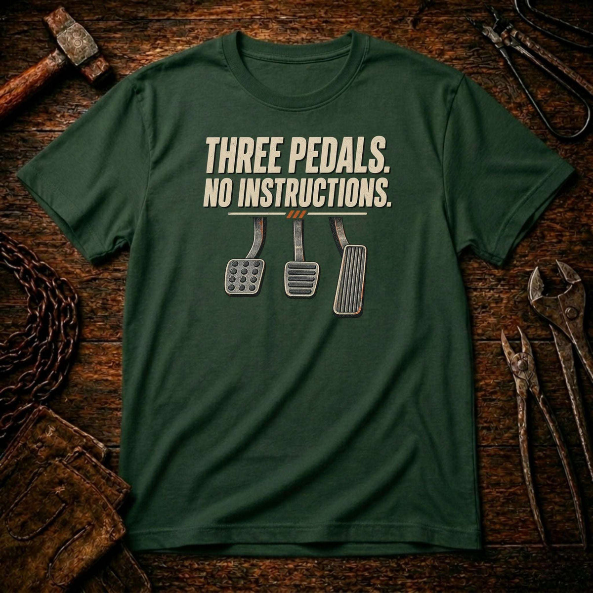 A dark green graphic t-shirt laid flat on a rustic wooden surface surrounded by tools. The shirt features the text 'THREE PEDALS.