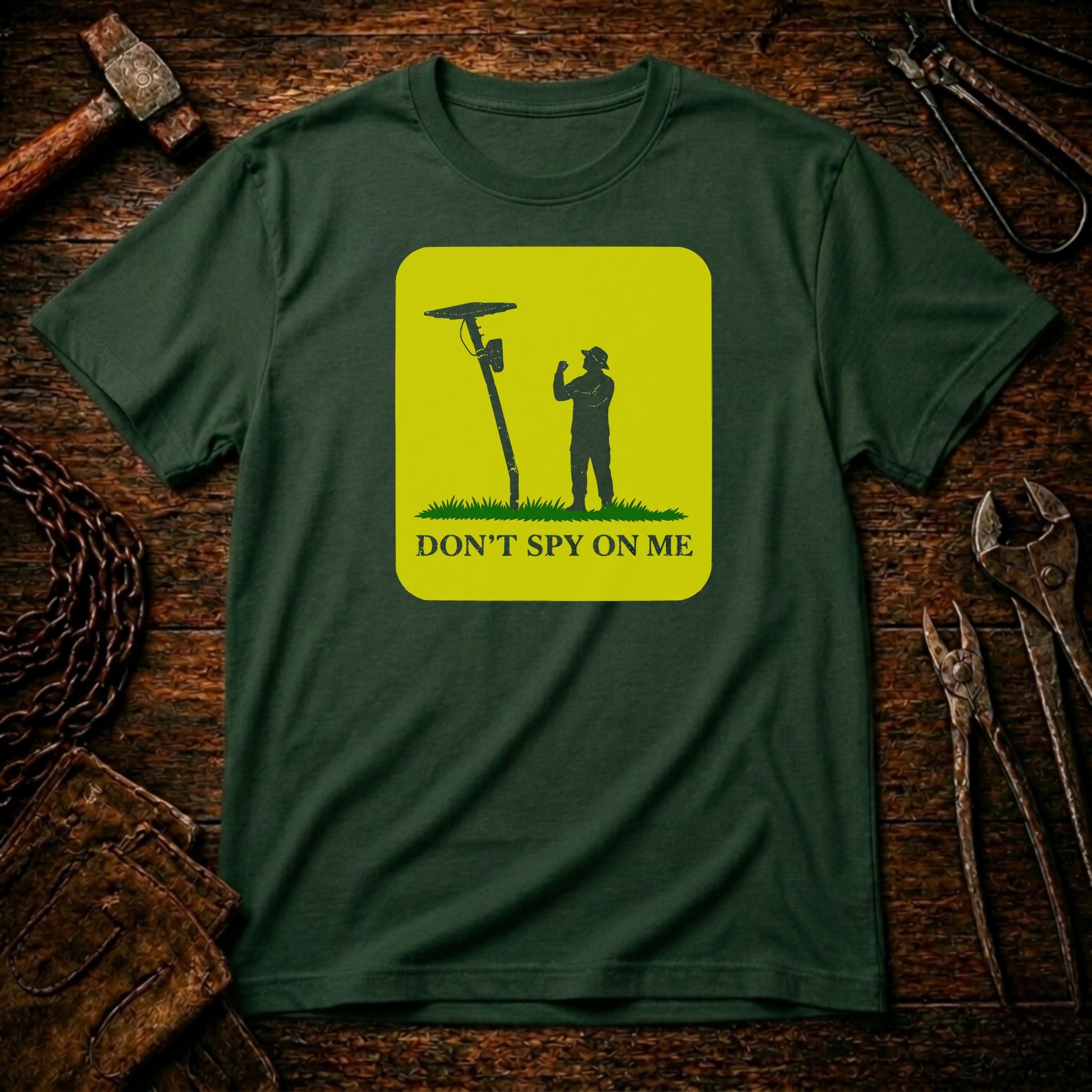 A dark green crewneck t-shirt with a bright yellow-green square graphic on the chest. The graphic shows a silhouette of a person holding a spy camera next to a lamp post, with the text 'DON'T SPY ON ME' below.